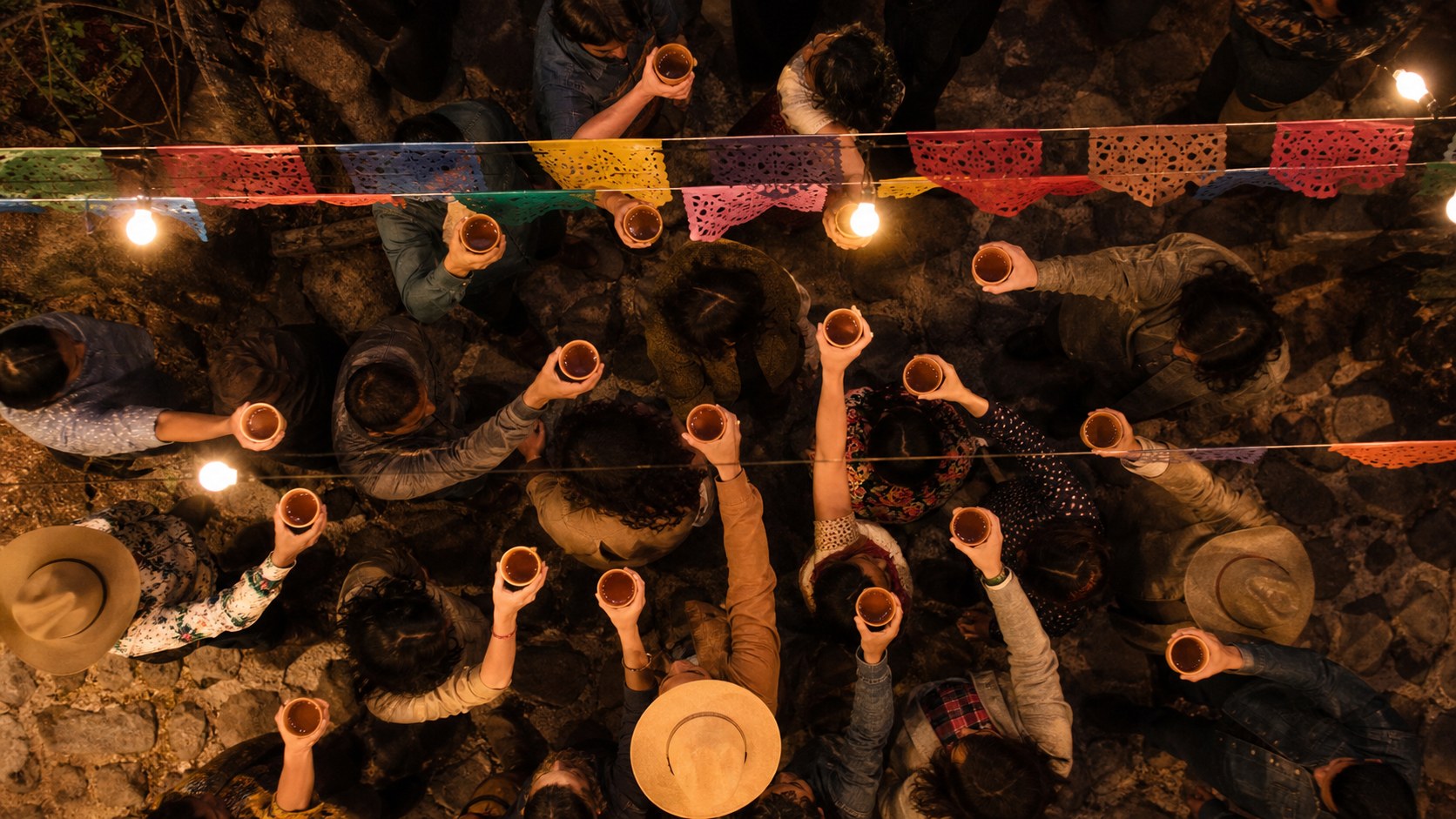 A crowded courtyard at night from above, a dozen arms raised at the same instant with copitas in them, everyone caught in the same gesture.