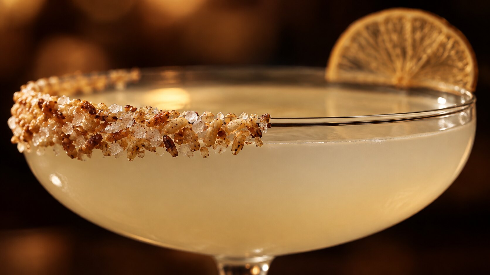 Extreme close-up of a coupe glass rim with a crust of toasted rice and coarse salt down one side and a slice of dehydrated citrus balanced on the far edge, the pale drink glowing in warm bar light.